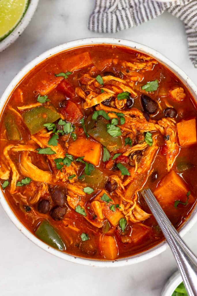 Chicken soup with sweet potatoes, black beans, and veggies garnished with in a bowl with a spoon in it. Around the bowl is a striped kitchen towel, a plate of lime wedges, and a bowl of cilantro.