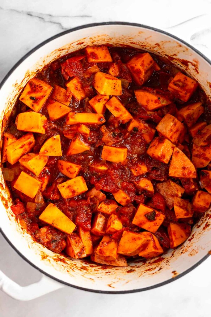 A mixture of diced sweet potatoes, canned tomatoes, onions, and spices in a white dutch oven.