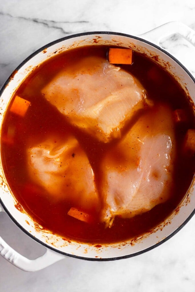 Raw chicken breasts overtop veggies covered with broth in white dutch oven.