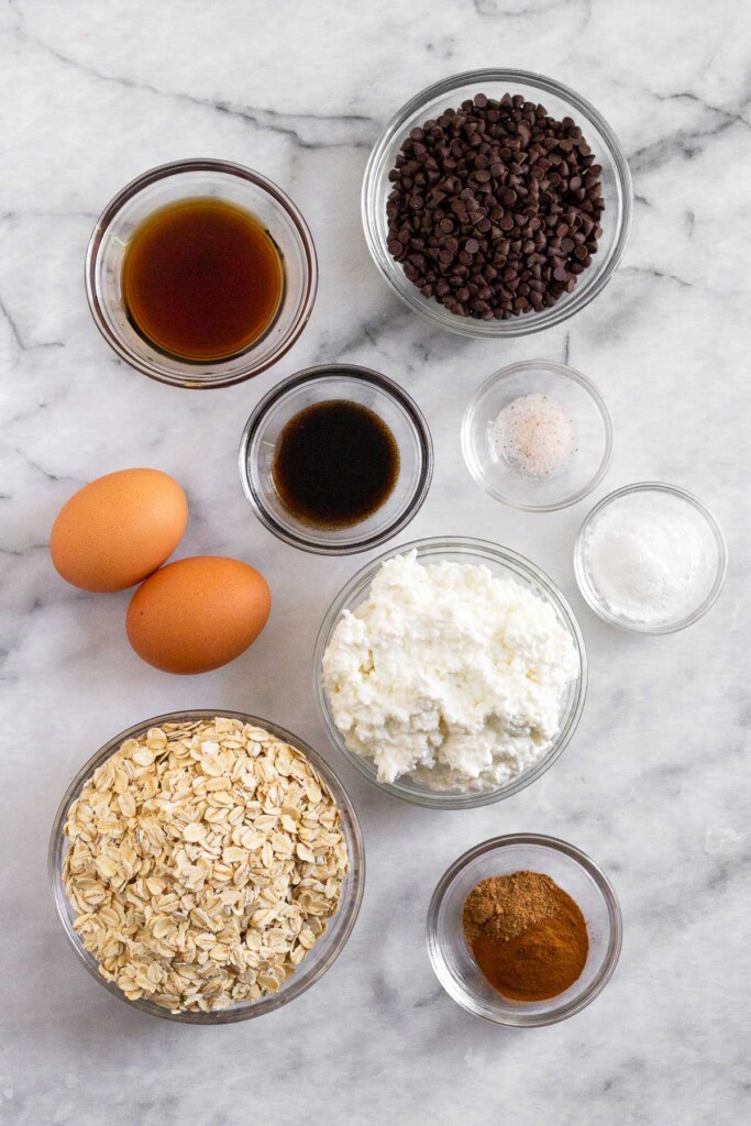 White marble counter top with a bowl of mini chocolate chips, a bowl of salt, a bowl of baking powder, a bowl of cottage cheese, a bowl of spices, a bowl of rolled oats, 2 brown eggs, a bowl of vanilla extract, and a bowl of maple syrup.