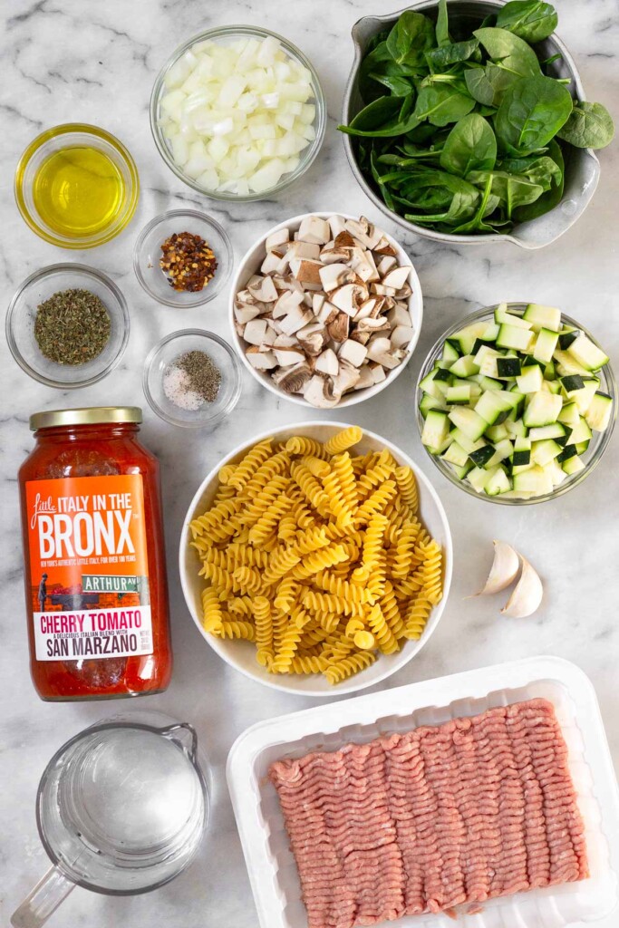 White marble counter top with a bowl of diced onion, a bowl of spinach, a bowl of diced mushrooms, a bowl of diced zucchini, 2 garlic cloves, a bowl of fusilli, a container of ground turkey, a cup of water, a jar of pasta sauce, a small bowl of salt and pepper, a bowl of Italian seasoning, a bowl of red pepper flakes, and a bowl of olive oil.