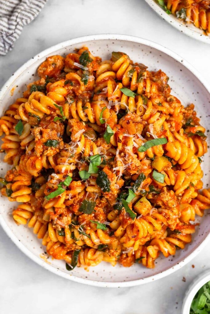 Overhead shot of pasta with ground turkey and veggies on a white plate garnished with parmesan cheese, fresh basil, and red pepper flakes. Off the the side is another plate of pasta and a small bowl of chopped basil.