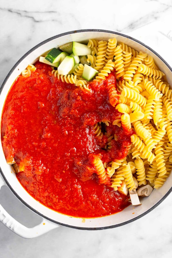 Large white dutch oven filled with uncooked pasta, veggies, pasta sauce, and ground meat.