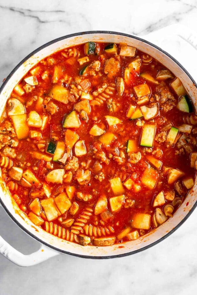 Large white dutch oven filled with ground meat, diced veggies, pasta, and pasta sauce before it is cooked.