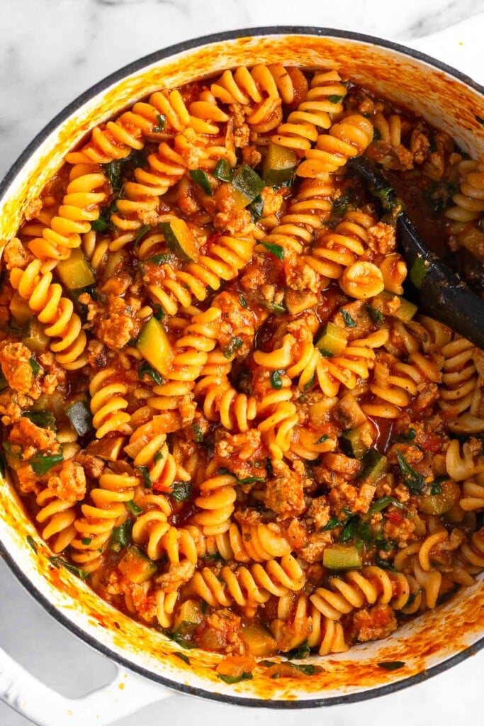 One pot pasta with meat and veggies in a large white dutch oven. There is a large spoon in the pot as well.