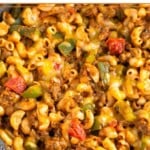 High protein ground beef recipe with pasta, bell peppers, and melted cheese in a large skillet. The skillet is on a striped towel and off to the side is a dish of cilantro.
