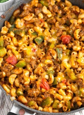 High protein ground beef recipe with pasta, bell peppers, and melted cheese in a large skillet. The skillet is on a striped towel and off to the side is a dish of cilantro.