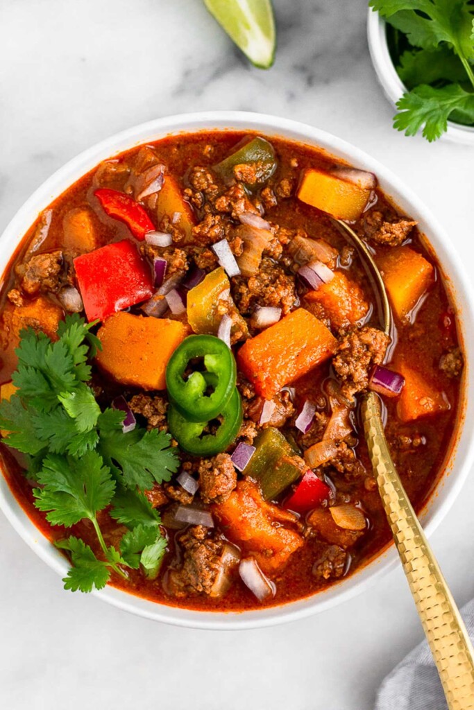 Bison chili with butternut squash and veggies in a bowl with a spoon in it. It is garnished with sliced a jalapeños and cilantro. Around it is a bowl of cilantro and lime wedges.