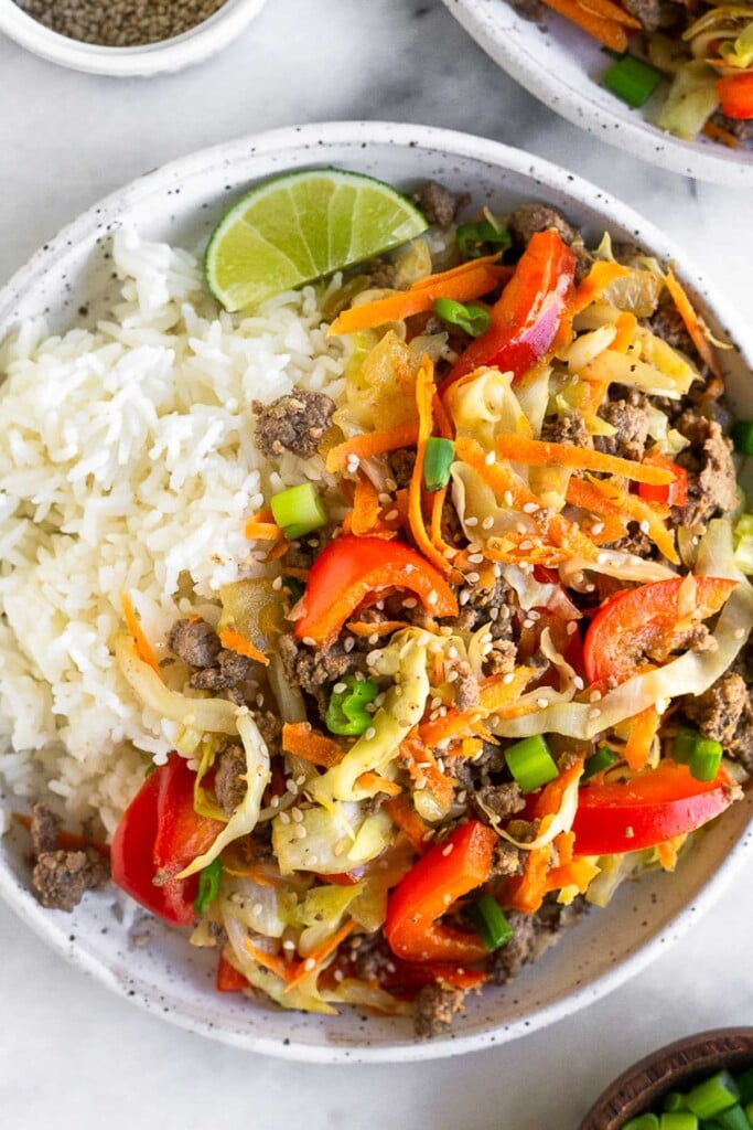 Highi protein ground beef stir fry recipe with cabbage, bell peppers, and carrots on a plate next to some white rice. It is garnished with a lime wedge, green onions, and sesame seeds.