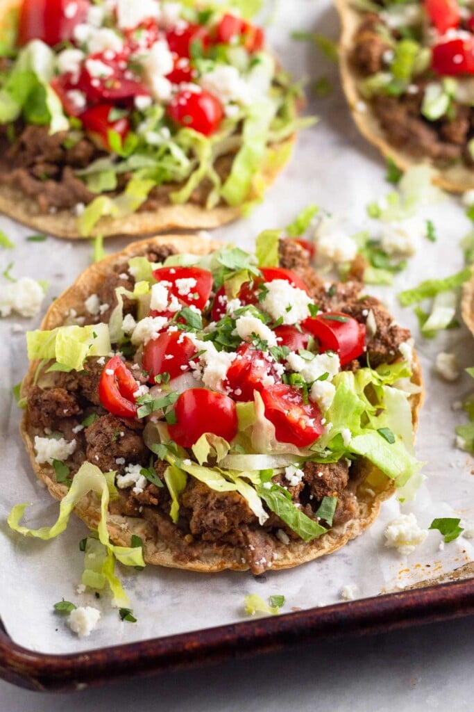 Beef tostada with beans, taco meat, lettuce, tomato, cheese, and cilantro. Behind it is another one and they are on a baking sheet lined with parchment.