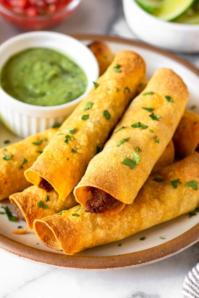 Beef and cheese taquitos garnished with cilantro stacked on top of each other on a plate. Also on the plate is a ramekin of avocado sauce. Behind the plate is a bowl of salsa and a bowl of lime wedges.