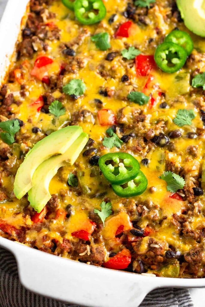 High protein ground beef casserole with rice, beans, bell peppers, and topped with melted cheese in a large casserole dish. It is garnished with avocado, jalapeño, and cilantro.