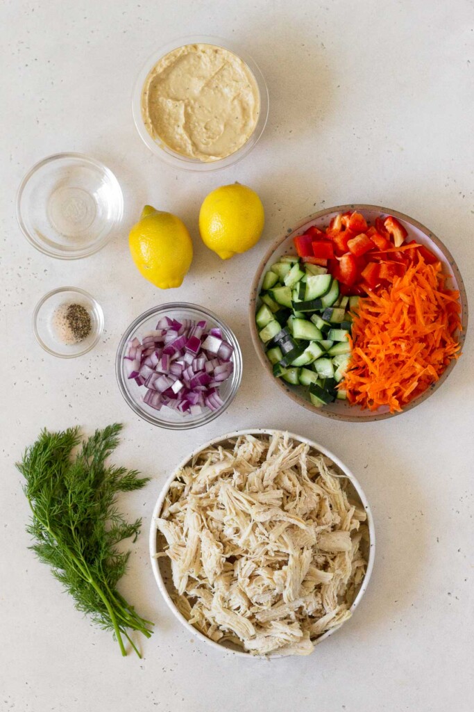 White countertop with a container of hummus, a bowl of chopped cucumber, chopped red bell peppers, and shredded carrots, a bowl of shredded chicken, a bunch of fresh dill, a bowl of diced red onions, a bowl of salt and pepper, two whole lemons, and a bowl of water.