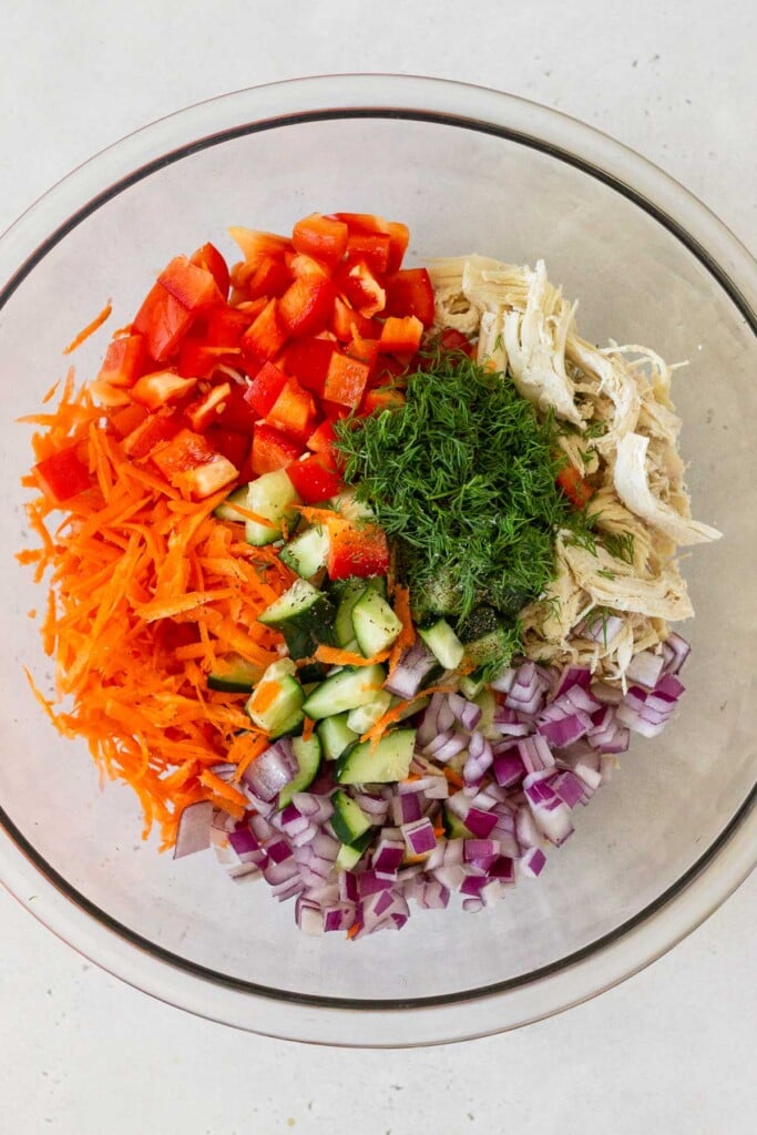 A large glass bowl of shredded chicken, diced red onion, shredded carrots, diced cucumber, diced red bell pepper, and fresh dill.
