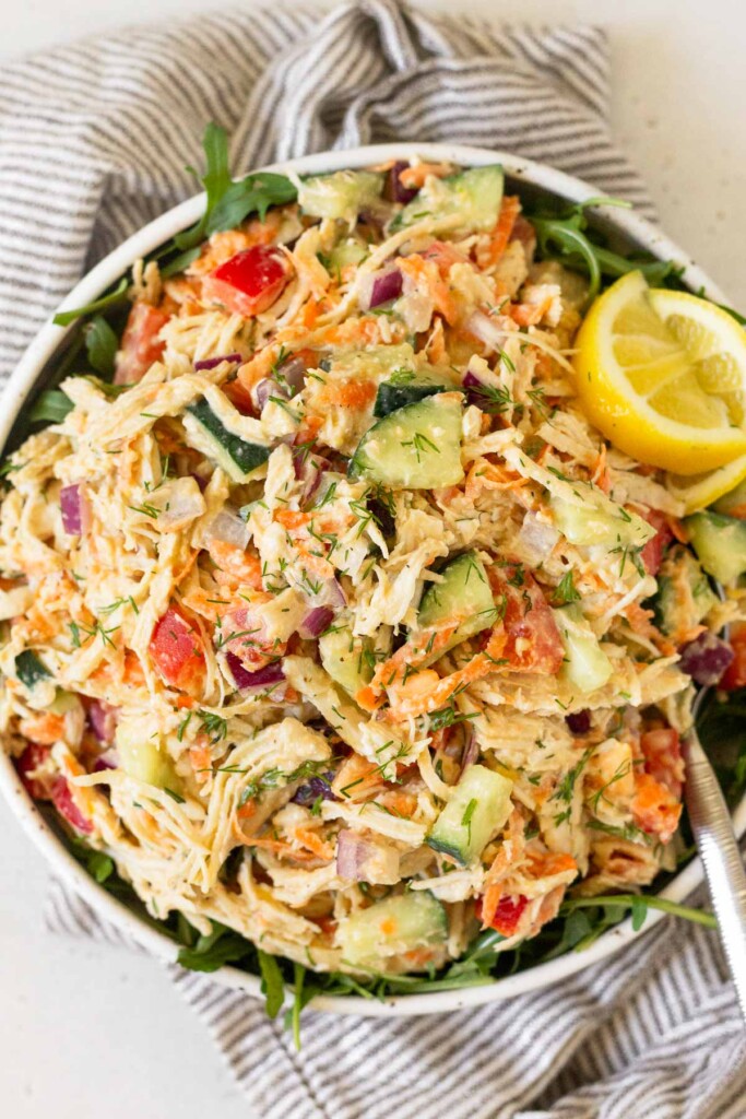 Chicken and hummus salad with peppers, carrots, cucumber, and red onion in a bowl overtop greens. The bowl is sitting on a striped kitchen towel.
