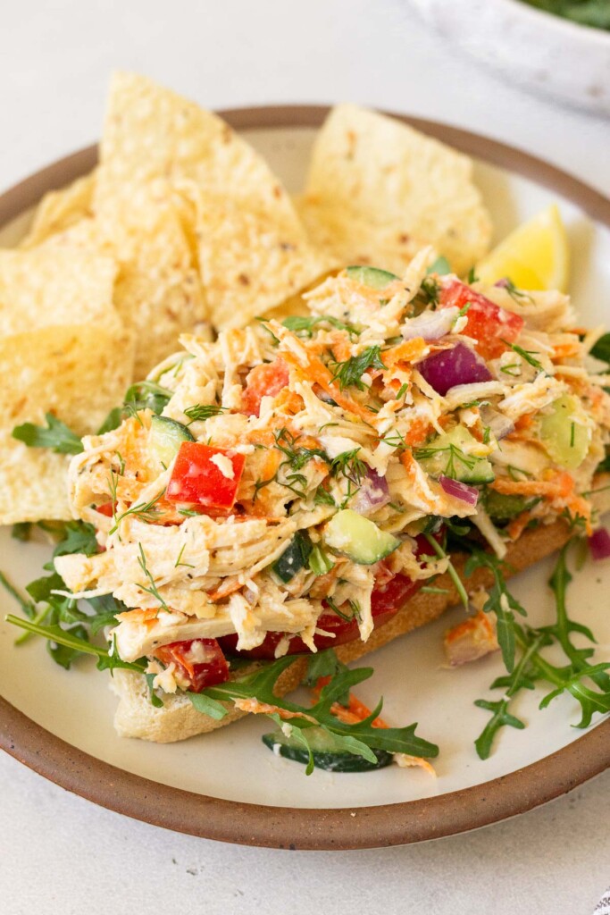 Chicken salad with hummus and veggies on top of some arugula on a piece of toast on a plate. Also on the plate are some tortilla chips.