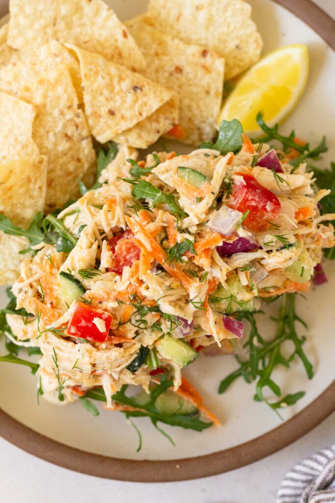 Chicken salad on top of some arugula on a piece of bread on a plate. There is also a lemon wedge and some tortilla chips on the plate.