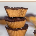 Three protein peanut butter cups stacked on top of each other on a white marble counter. The top one has a bite taken out of it. Around them are more PB cups.
