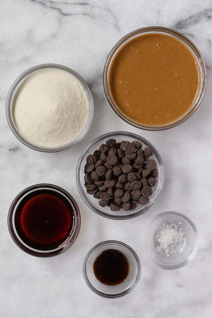White marble counter with a bowl of peanut butter, a bowl of chocolate chips, a bowl of sea salt, a bowl of vanilla extract, a bowl of maple syrup, and a bowl of collagen.