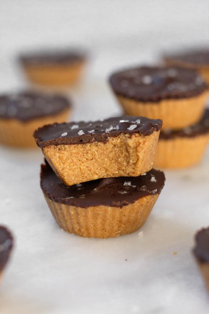 Two high protein peanut butter cups on a white marble counter. The top one has a bite taken out of it. Around them are more peanut butter cups.