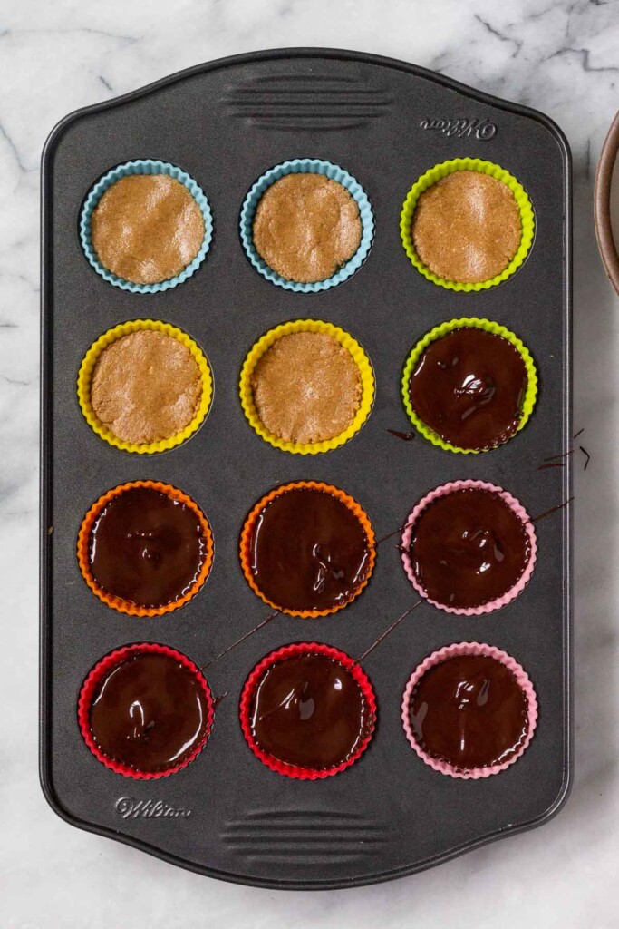 Mini muffin tin lined with silicone liners with a peanut butter dough in them. Half of them are covered in melted chocolate.