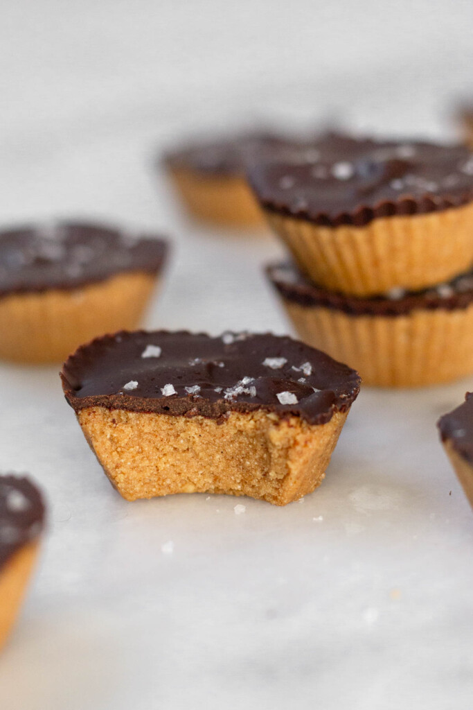 Protein Reese’s peanut butter cup with a bite taken out of it on a white marble counter. Around it are more peanut butter cups and two of them are stacked on top of each other.