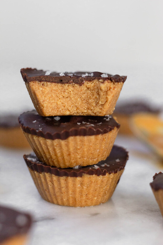 Three protein peanut butter cups stacked on top of each other on a white marble counter. The top one has a bite taken out of it. Around them are more PB cups.