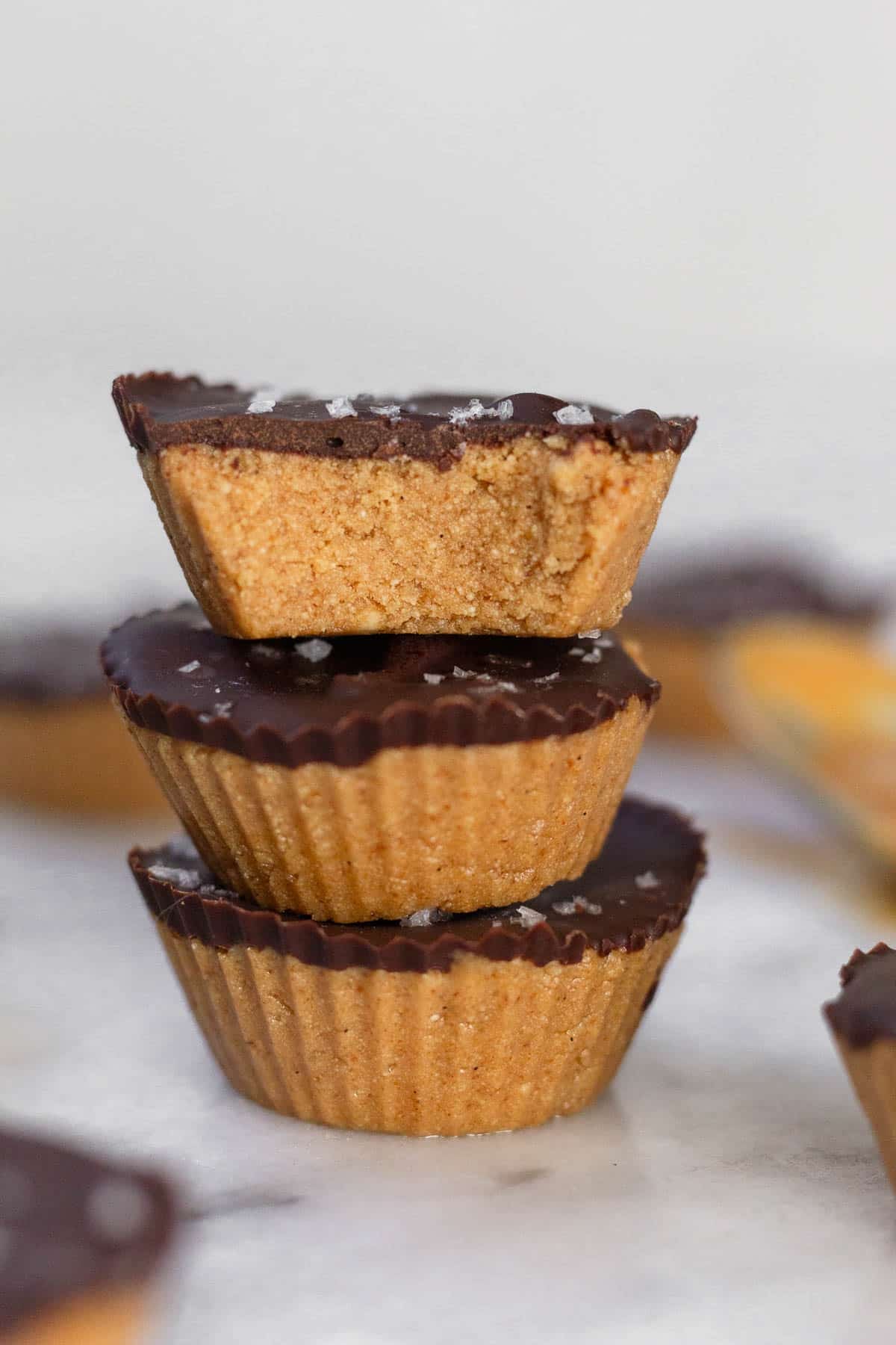 Three protein peanut butter cups stacked on top of each other on a white marble counter. The top one has a bite taken out of it. Around them are more PB cups.