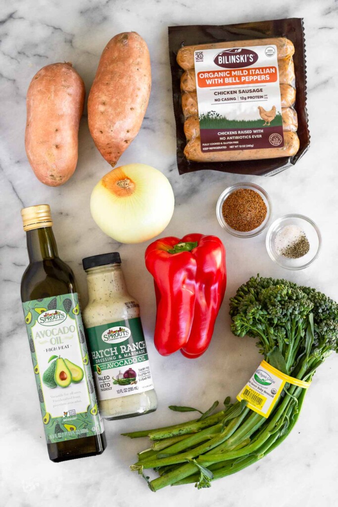 White marble counter with a package of chicken sausages, a bowl of spices, a small bowl of salt and pepper, a bunch of baby broccoli, a large red pepper, a bottle of ranch, a bottle of avocado oil, a yellow onion, and two sweet potatoes.