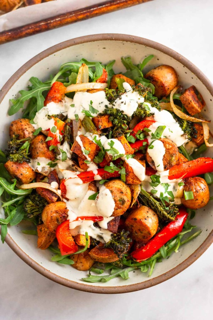 A bowl filled with arugula, roasted veggies, and roasted chicken sausage slices all drizzled with ranch and topped with fresh herbs. Behind it is a sheet pan.