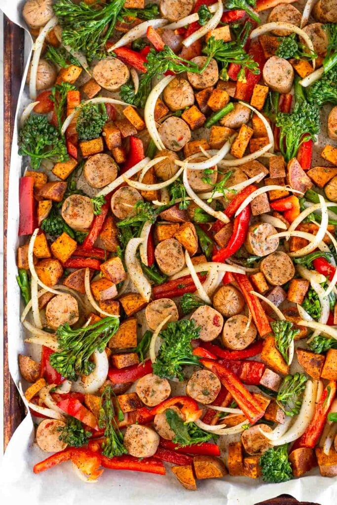 Large baking sheet filled with diced sweet potatoes, sliced onions, sliced peppers, broccoli, and sliced chicken sausages before they are roasted.