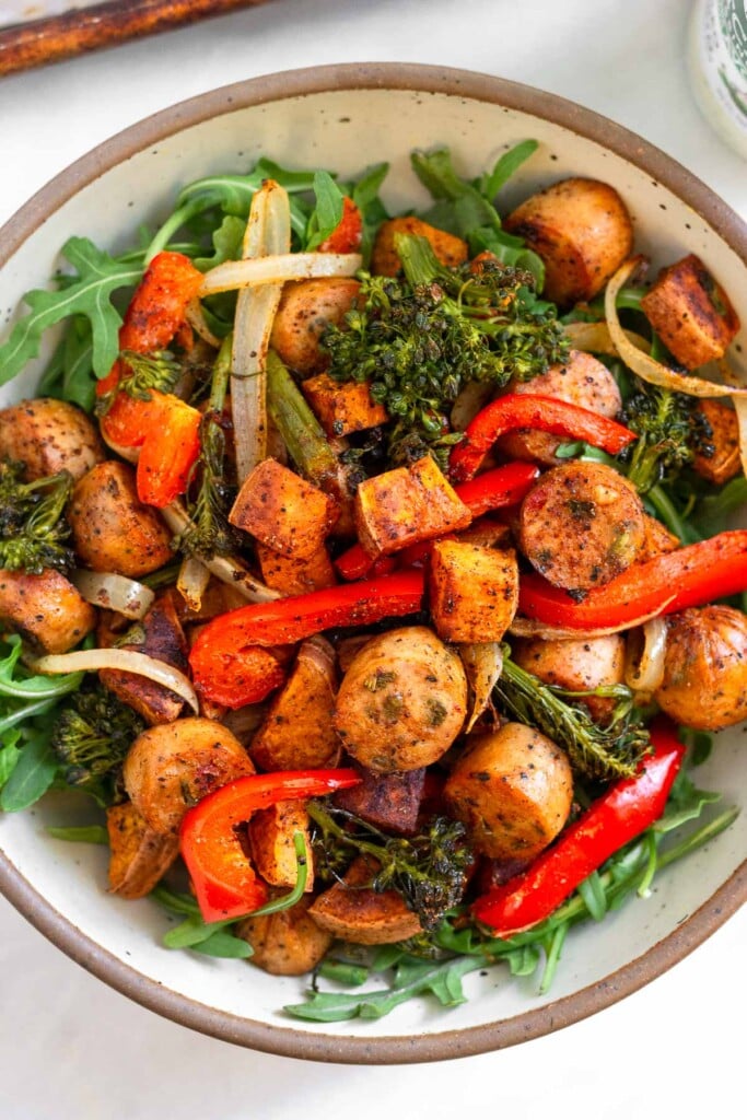 A white bowl filled with greens, roasted veggies, and sliced roasted chicken sausages.