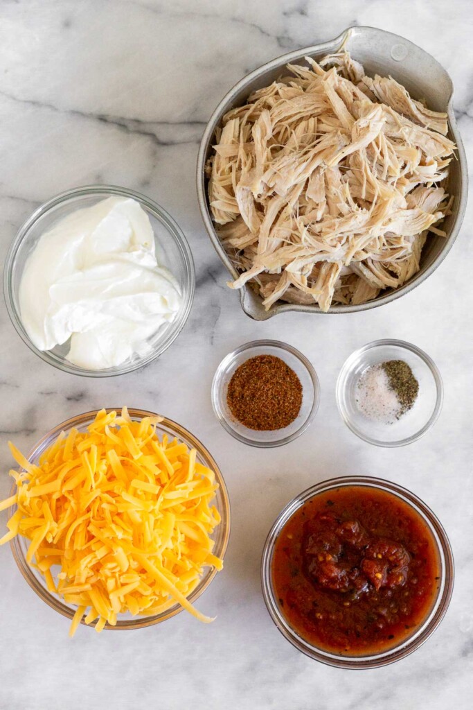 White marble counter with a bowl of shredded chicken, a small bowl of salt and pepper, a small bowl of taco seasoning, a bowl of red salsa, a bowl of shredded orange cheese, and a bowl of yogurt.