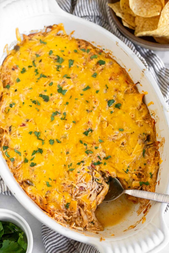 Shredded chicken dip topped with melted cheese and garnished with cilantro in a white baking dish. There is a spoon in the dish with seam of the dip missing. Next to the dish is a small bowl of cilantro and a bowl of tortilla chips.