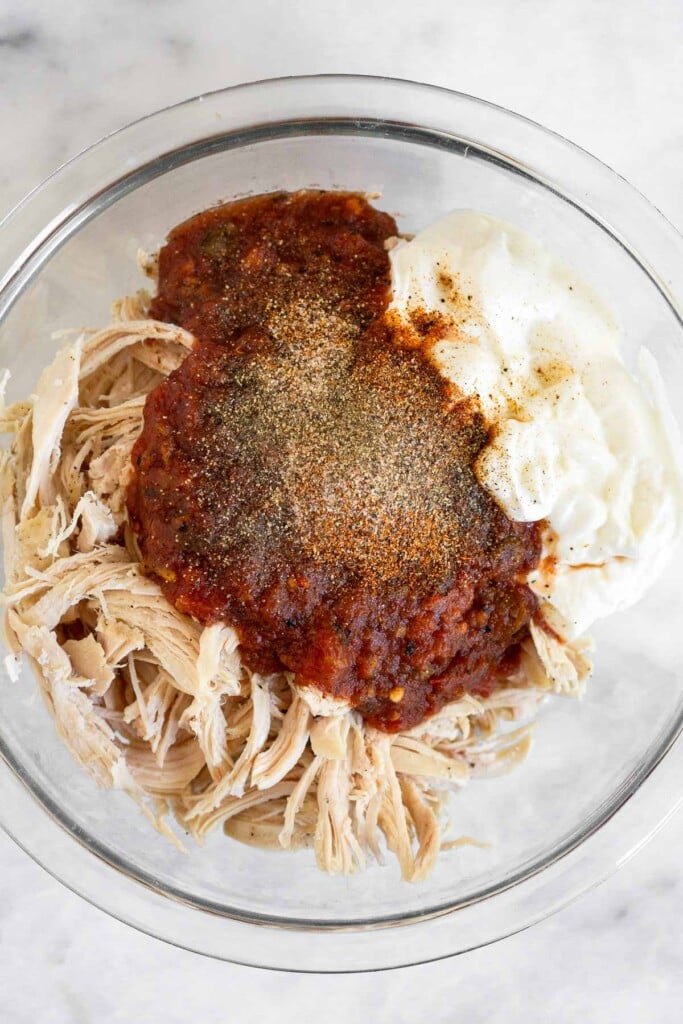Glass bowl of shredded chicken, greek yogurt, salsa, and seasoning before it is mixed together.