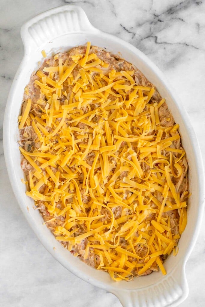 White baking dish with chicken dip spread across the Botton and topped with shredded cheese before it is baked.