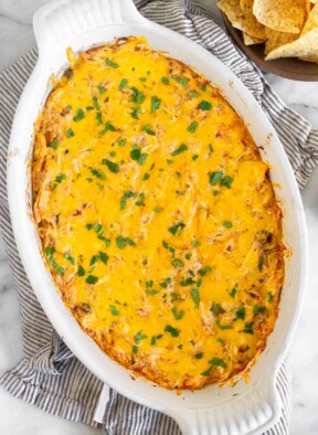 White baking dish with cheesy chicken dip garnished with cilantro. The dish is on a striped kitchen towel with a bowl of tortilla chips next to it.