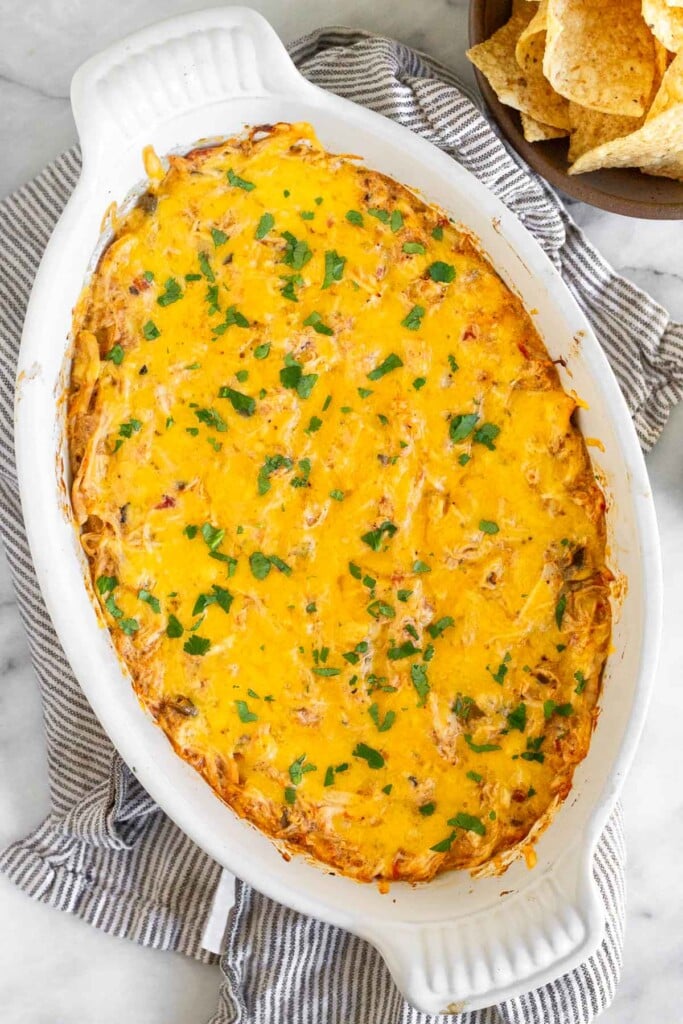 White baking dish with cheesy chicken dip garnished with cilantro. The dish is on a striped kitchen towel with a bowl of tortilla chips next to it.