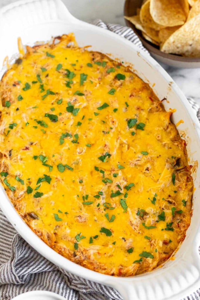 Hot chicken dip topped with melted cheese and cilantro in a white baking dish. Behind the dish is a bowl of tortilla chips.