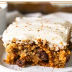 Carrot cake bar topped with frosting and cinnamon on a small plate. Behind it is the pan of the rest of the bars.