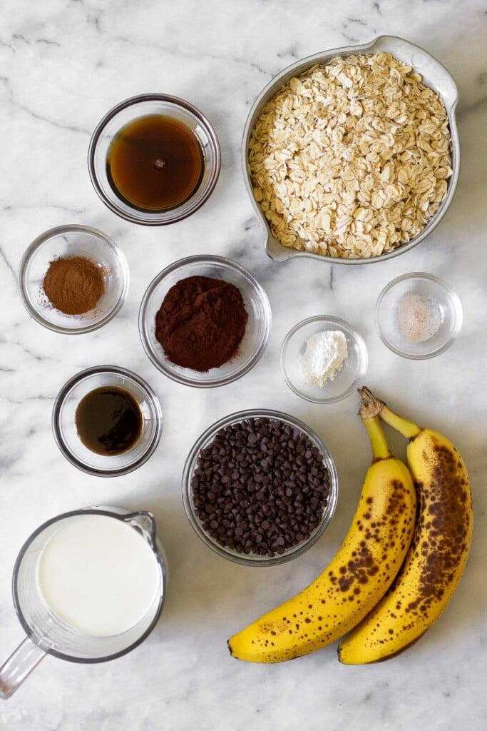 White marble counter with a bowl of rolled oats, a bowl of salt, a bowl of baking powder, 2 spotted bananas, a bowl of chocolate chips, a measuring cup of milk, a bowl of vanilla extract, a bowl of cocoa powder, a bowl of dinner, and a bowl of maple syrup.