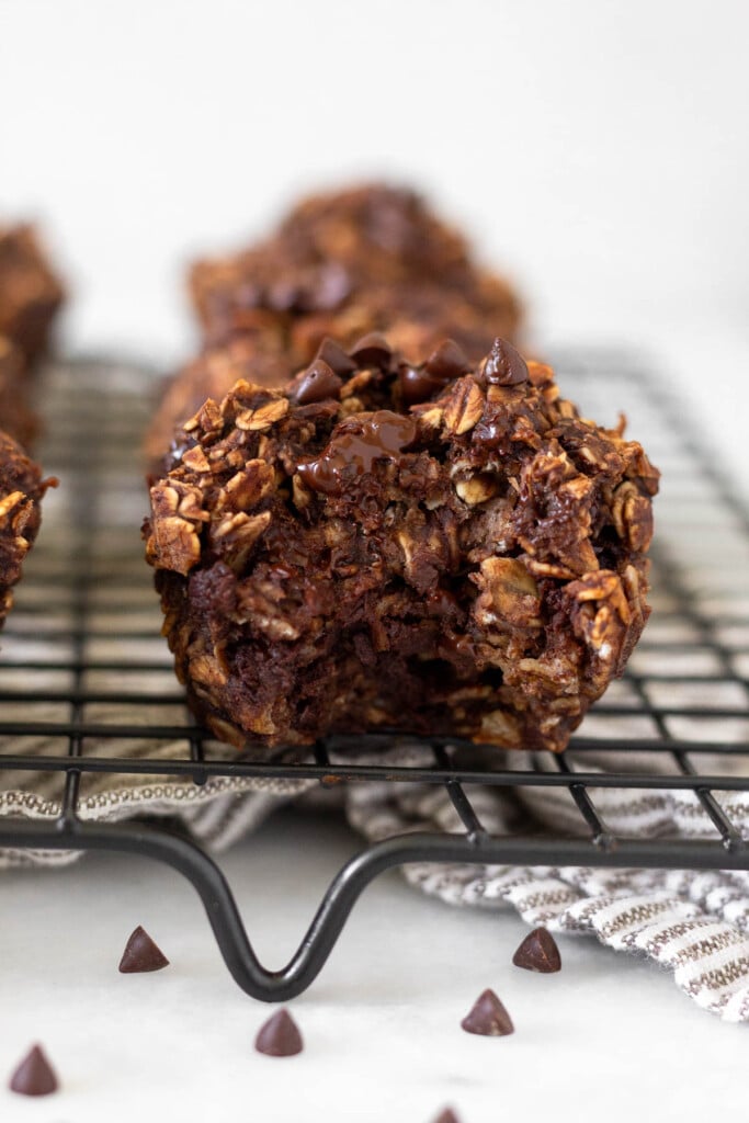 Cookie drying rack filled with banana chocolate oatmeal muffins with the one in the front having a bite taken out of it. In front of the rack are mini chocolate chips.
