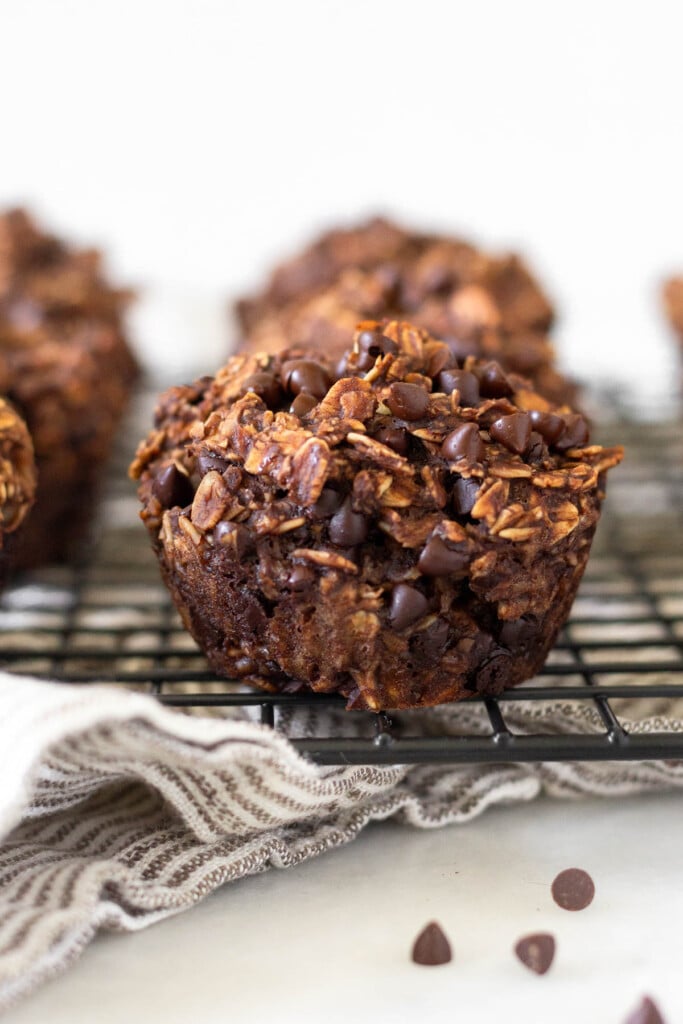 Close up of a chocolate banana oatmeal muffin on a cooling rack. There are more muffins surrounding it and some mini chocolate chips in front of it.