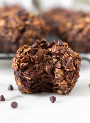 Chocolate oatmeal muffin with a bite taken out of it with some mini chocolate chips around it. Behind it is a rack of more muffins.