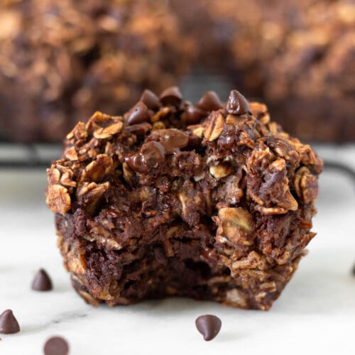 Chocolate oatmeal muffin with a bite taken out of it with some mini chocolate chips around it. Behind it is a rack of more muffins.