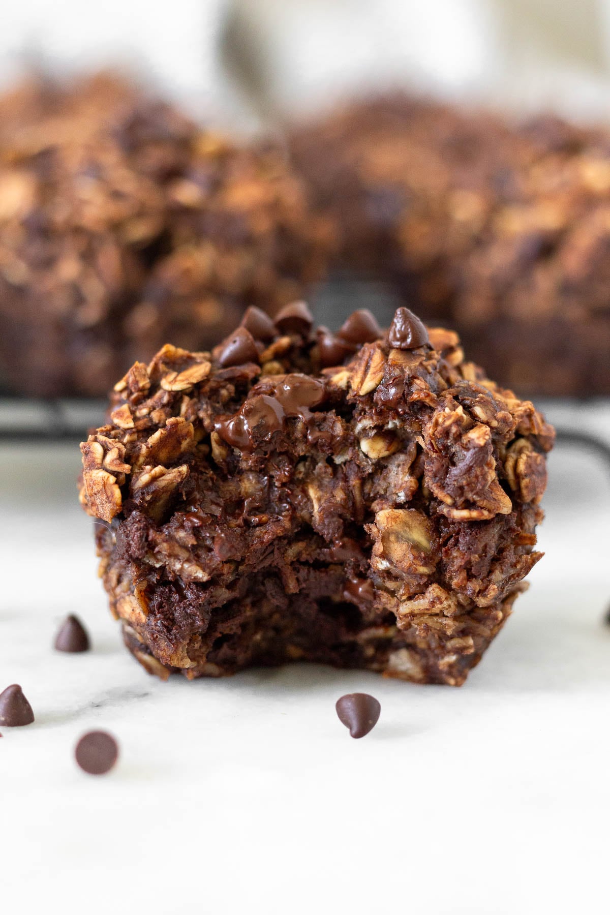 Chocolate oatmeal muffin with a bite taken out of it with some mini chocolate chips around it. Behind it is a rack of more muffins.