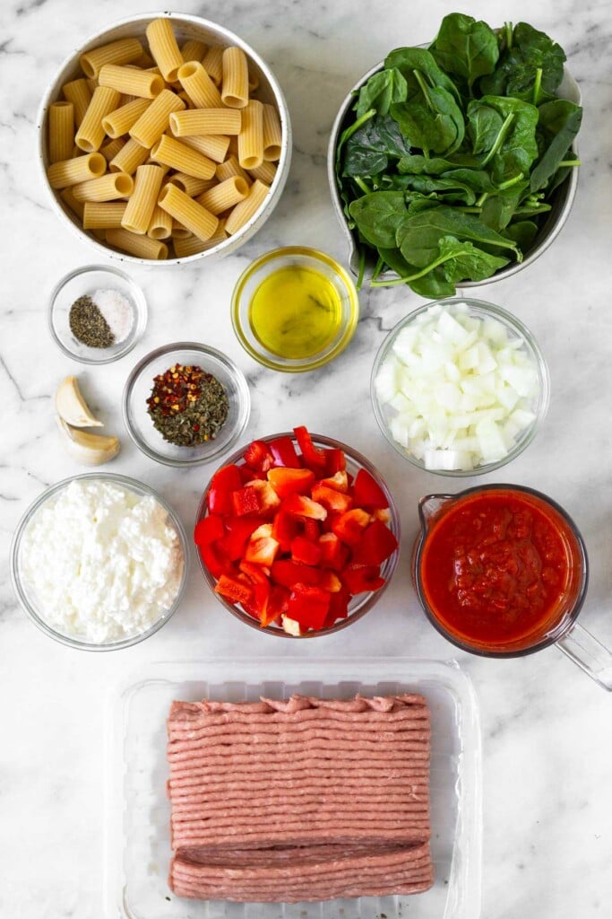 White marble counter with a bowl of spinach, a bowl of diced onion, a measuring cup of pasta sauce, a container of raw ground turkey, a bowl of diced bell pepper, a bowl of cottage cheese, a bowl of spices, 2 cloves of garlic, a bowl of salt and pepper, a bowl of olive oil, and a bowl of pasta.