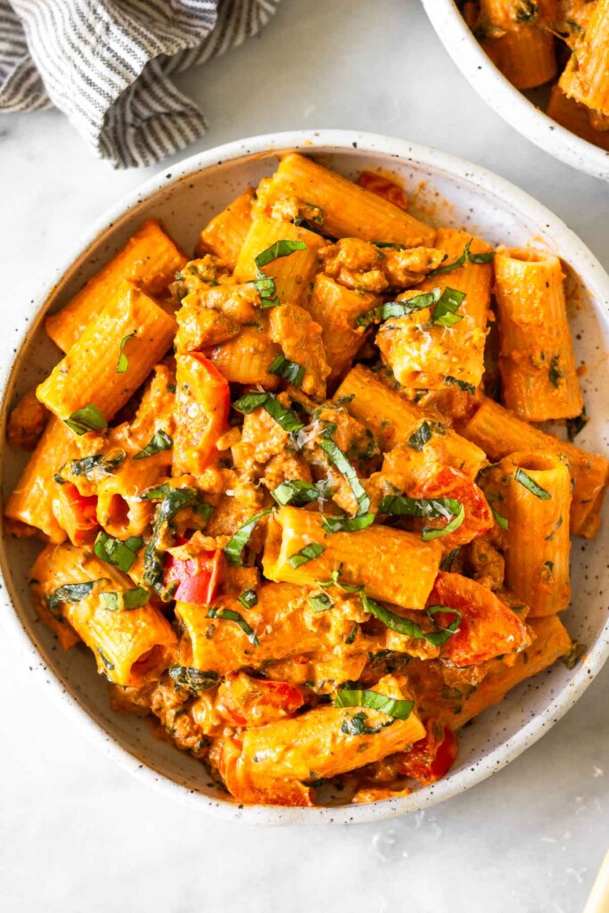 Bowl of pasta with ground meat and veggies tossed in a creamy marinara sauce. It is garnished with basil and parmesan cheese. Next to the plate is a striped kitchen towel and another bowl of pasta.