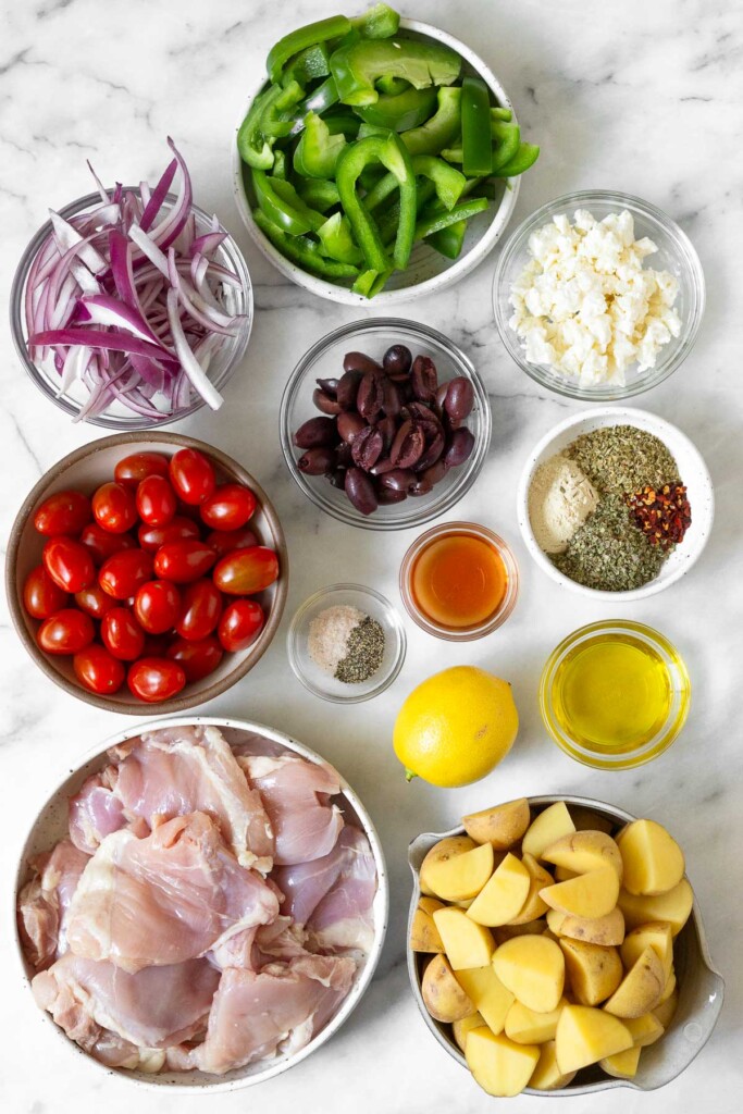 White marble counter with a bowl of sliced green pepper, a bowl of feta cheese, a bowl of spices, a bowl of olive oil, a bowl of quartered yellow potatoes, a bowl of raw chicken thighs, a whole lemon, a bowl of salt and pepper, and bowl of red wine vinegar, a bowl of cherry tomatoes, a bowl of halved Kalamata olives, and a bowl of sliced red onion.