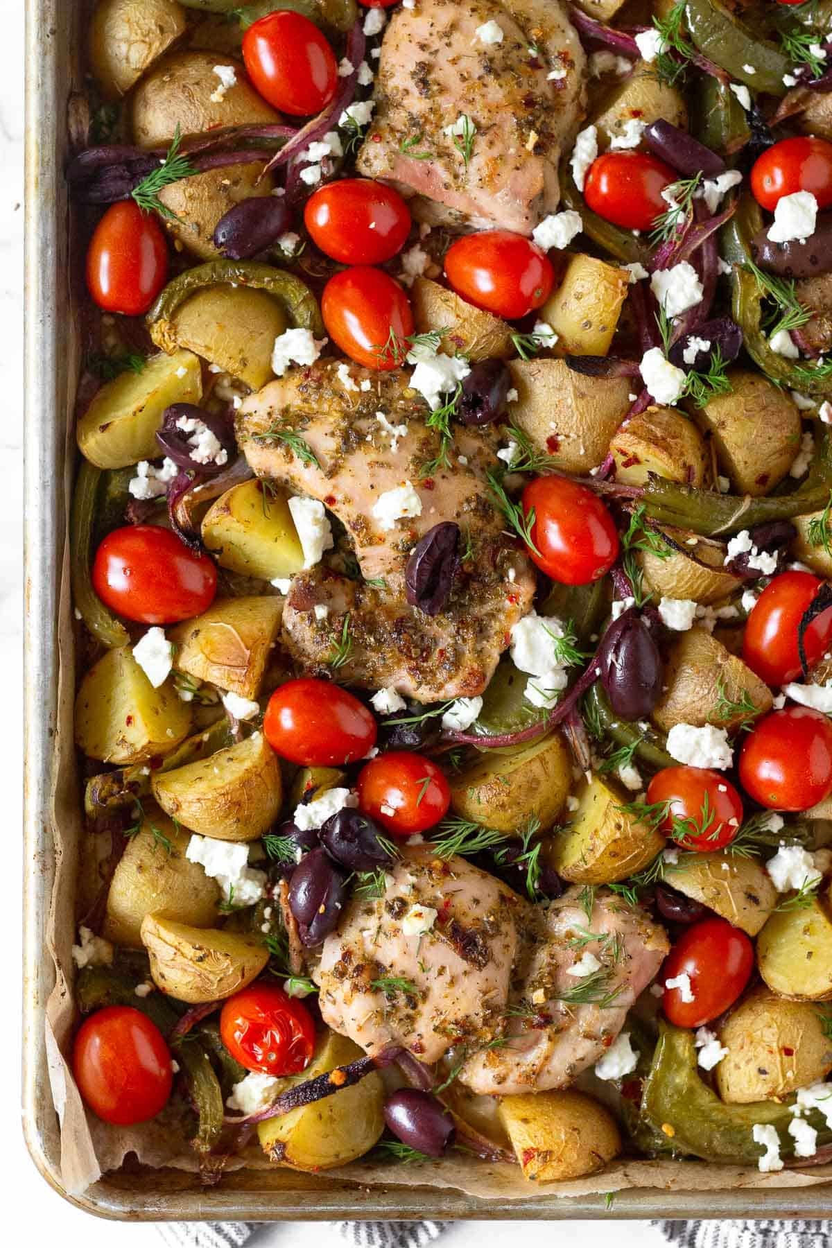 Greek sheet pan chicken with roasted potatoes, peppers, and onions, cherry tomatoes, Kalamata olives, and feta. It is garnished with fresh dill.